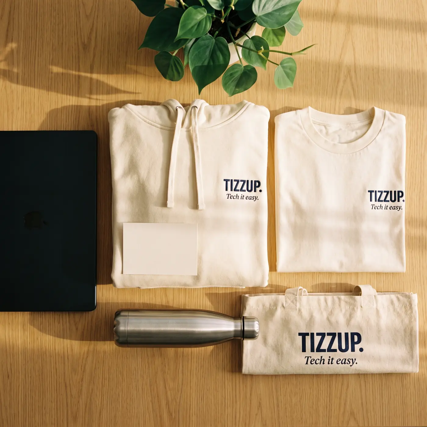 TIZZUP's company welcome pack laid out on a desk: ecru hoodie, t-shirt and tote bag marked in navy, a stainless steel bottle and a welcome card.