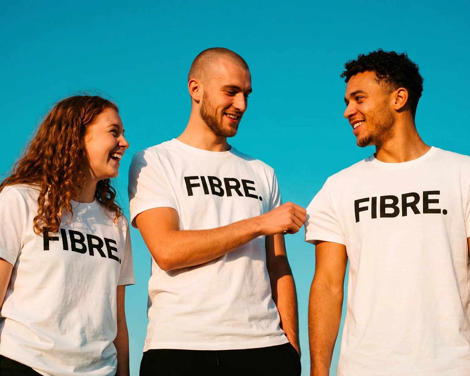 Three friends in white t-shirts printed with the word FIBRE laugh under a blue sky, one pinching his friend's sleeve to feel the combed cotton.