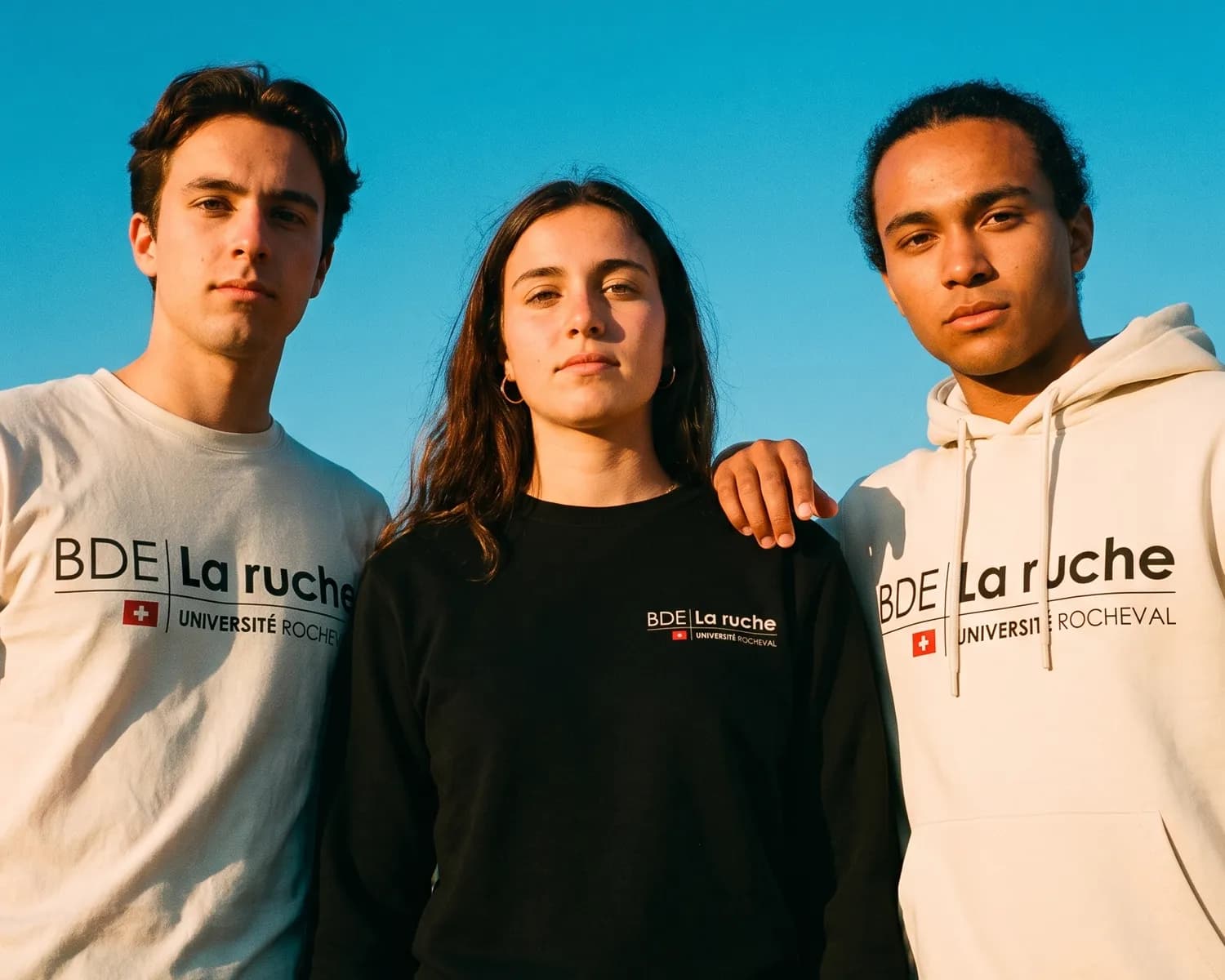 Three BDE La ruche students each wear their own size of their student union's custom clothing: an ecru t-shirt, a black sweatshirt and an ecru hoodie.