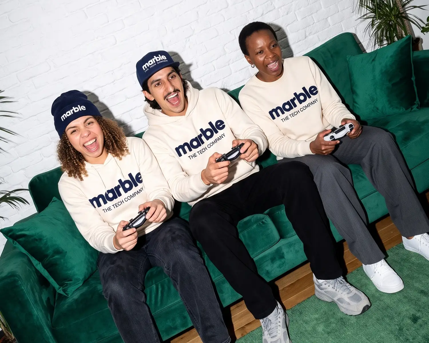 Three colleagues wear their company's seminar merch, ecru sweatshirts and navy beanies branded marble, mid video game on a green sofa.