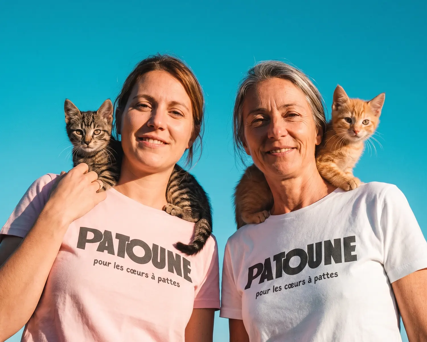 Deux bénévoles de l'association PATOUNE, en t-shirt personnalisé rose et blanc imprimé en gris anthracite, posent avec un chaton sur l'épaule devant un ciel bleu : le t-shirt vendu pour financer une association.
