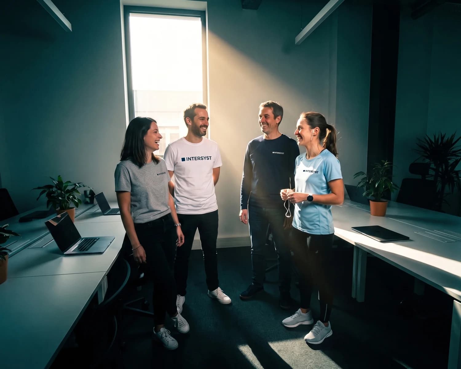 Quatre collègues de l'entreprise INTERSYST discutent dans leur open space, chacun dans un t-shirt personnalisé différent au logo de l'entreprise, dont une collègue en t-shirt d'entraînement bleu ciel prête à partir courir.
