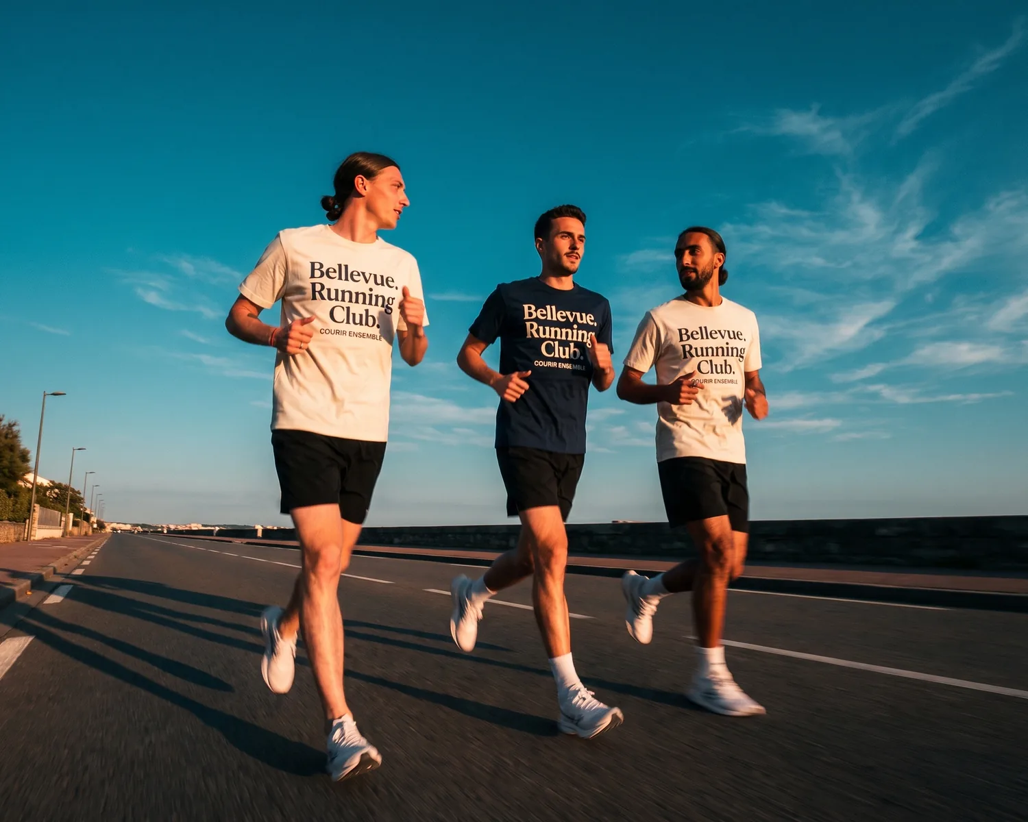 Trois coureurs du Bellevue Running Club courent ensemble sur une route du bord de mer au coucher du soleil, en t-shirts running personnalisés crème et marine au logo du club sur la poitrine.