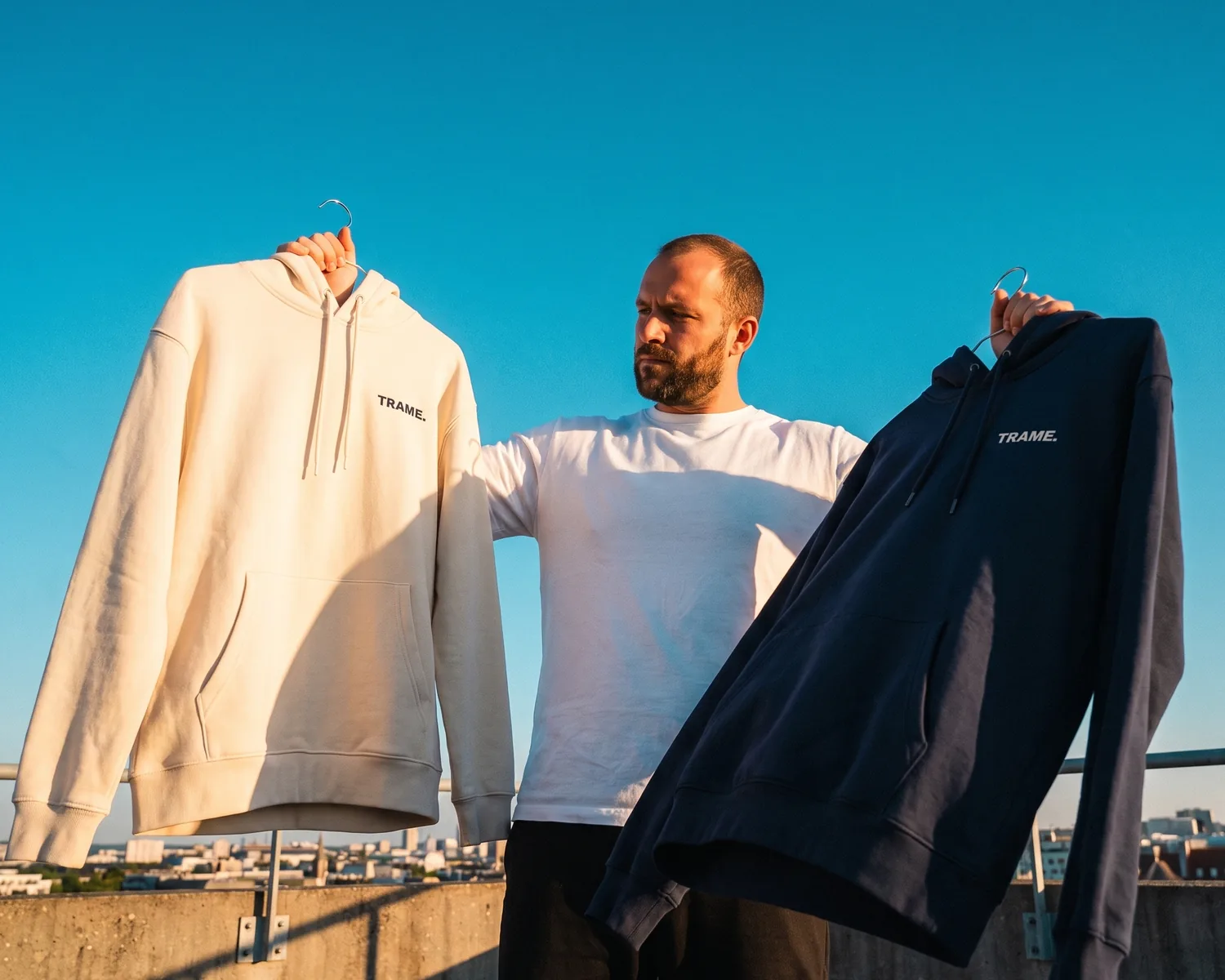 On a rooftop, a bearded man holds up two custom hoodies bearing the TRAME. logo: a heavy ecru hoodie that hangs straight and a lighter navy hoodie that folds softly.