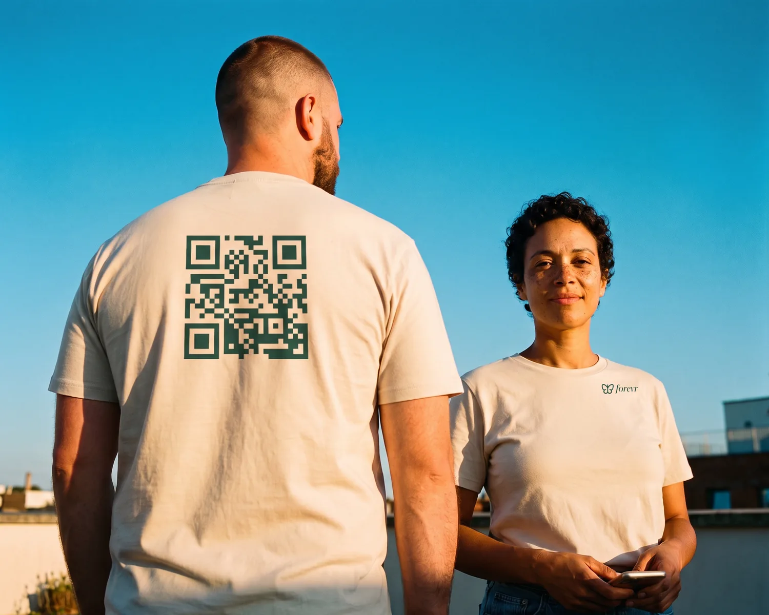 On a rooftop, a man seen from behind wears an ecru t-shirt with a large green QR code printed on the back, facing a woman whose ecru t-shirt bears the forevr logo on the chest.