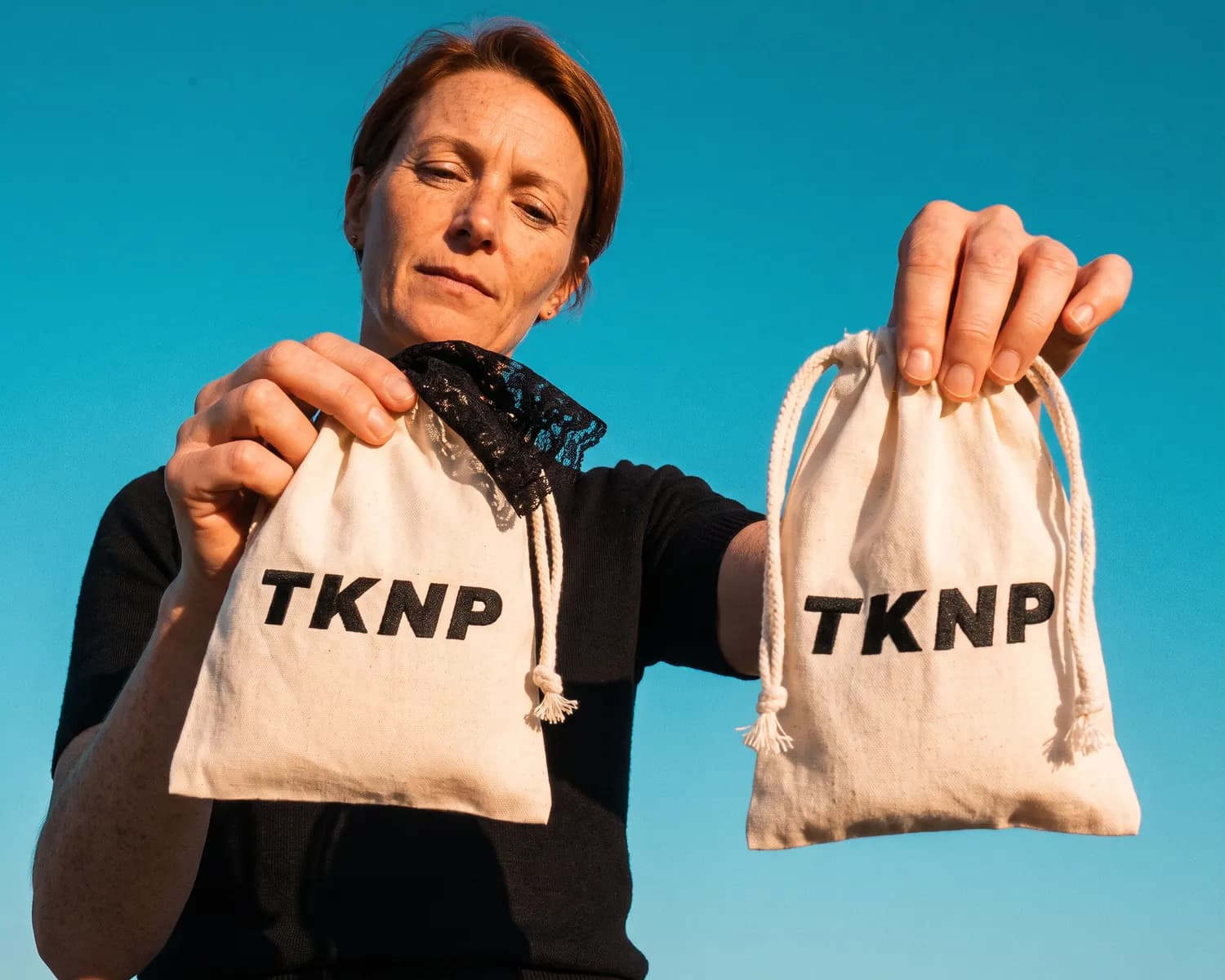 Under a blue sky, a woman holds out two custom drawstring pouches in ecru cotton, closed by a cord and marked with the TKNP logo in black; a piece of black lace peeks out of one of them.