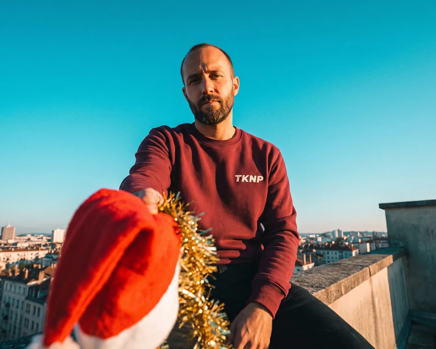 A man in a custom burgundy sweatshirt with the TKNP logo, sitting on his office rooftop, holds out toward the camera the Santa hat and tinsel from Christmas Jumper Day.