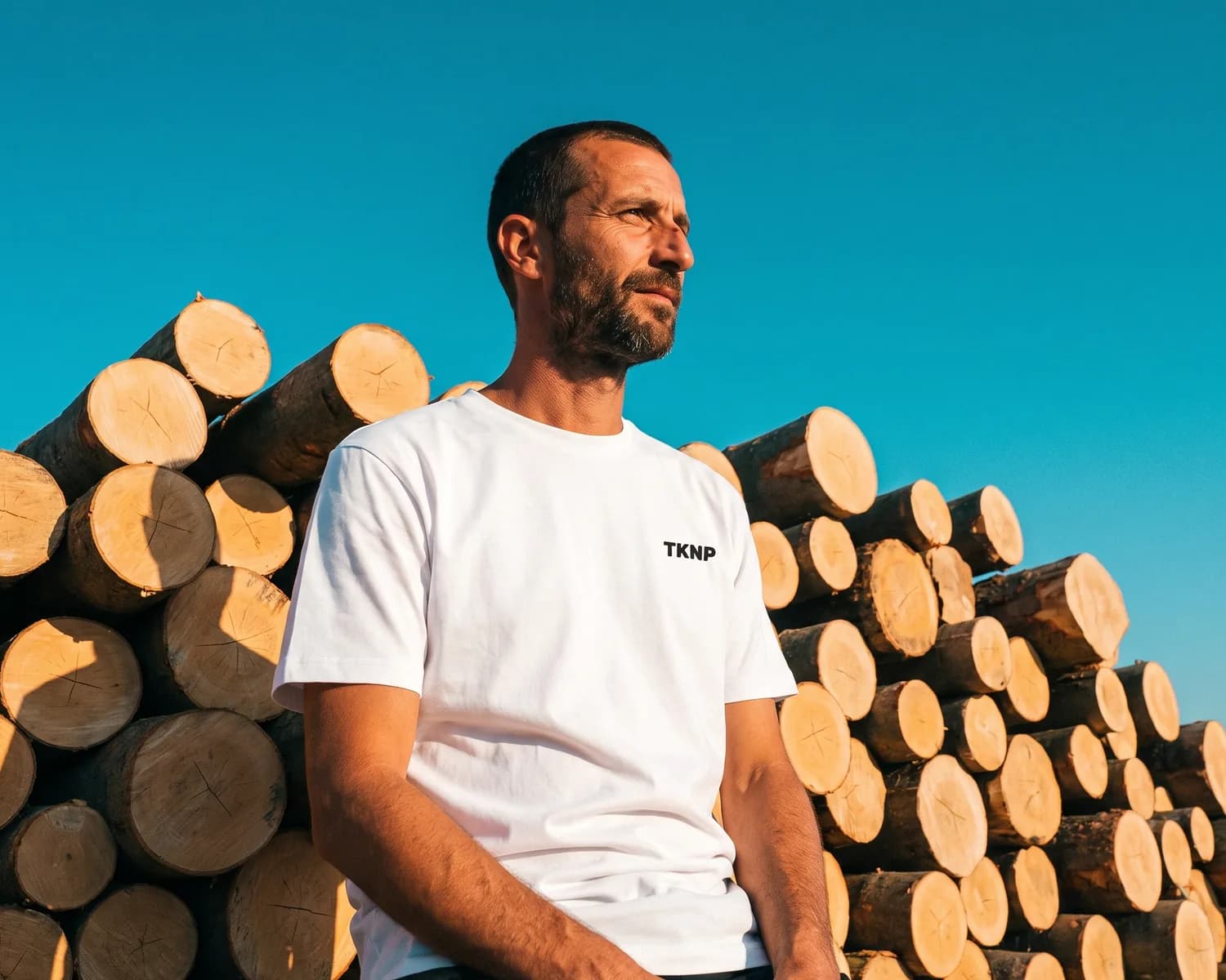 A man in a white modal t-shirt, a blend of organic cotton and modal, with the TKNP logo printed on the chest, sitting against a stack of logs: modal is a fibre made from wood.