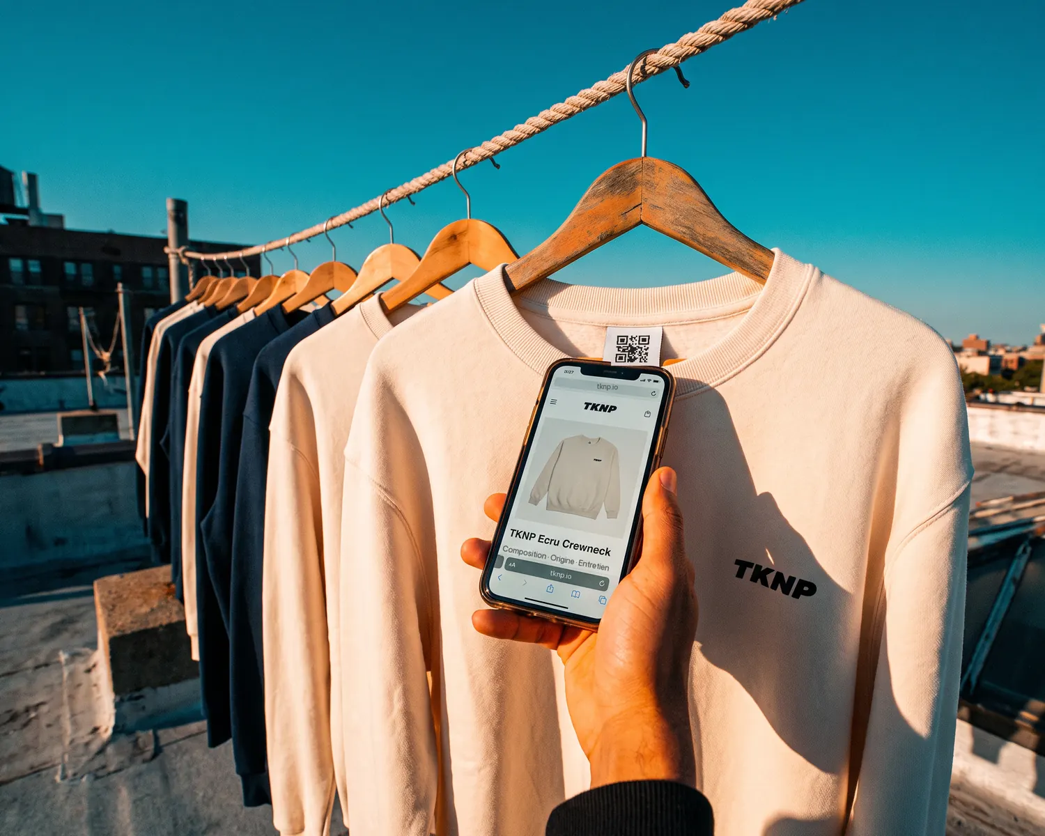 A hand scans with a phone the QR code on the neck label of an ecru TKNP sweatshirt, hanging with the ecru and navy collection: the digital product passport opens on the screen.
