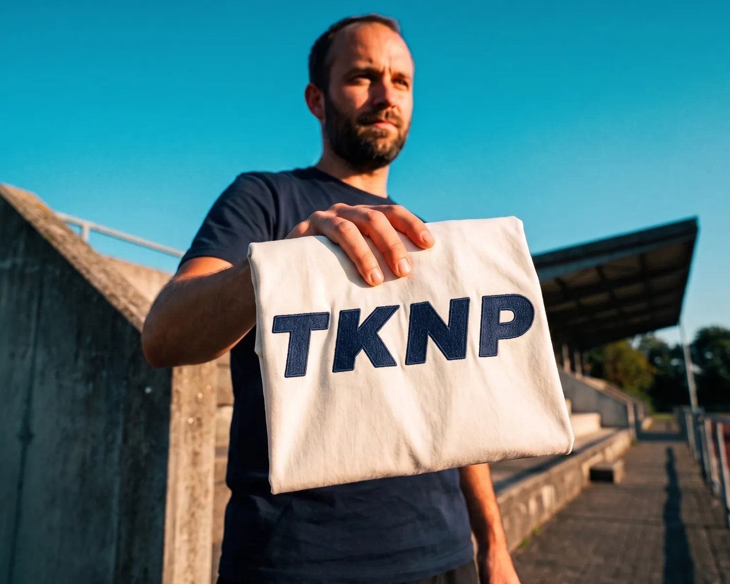 An association volunteer holds out a custom ecru t-shirt embroidered with the TKNP logo in navy thread, at the top of a sports ground stand.