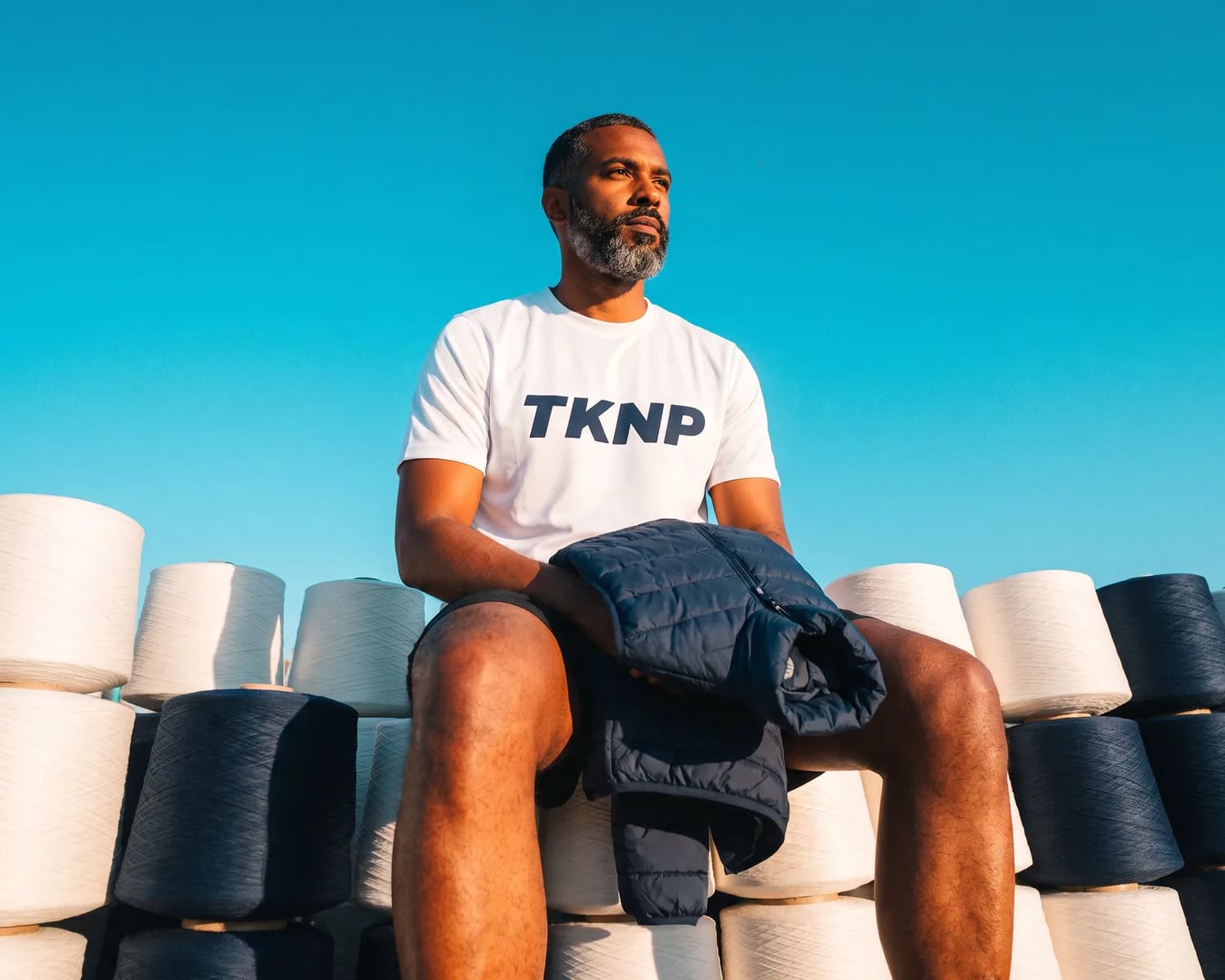 Fibre synthétique : un homme en t-shirt de sport blanc TKNP tient un gilet matelassé marine, assis sur des bobines de fil