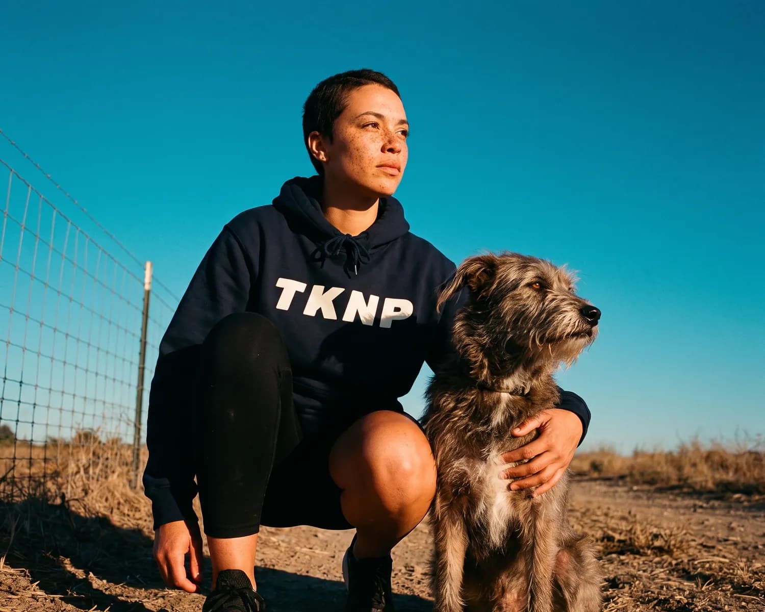 PETA-Approved Vegan label: a shelter volunteer in a navy TKNP hoodie, crouching by a wire fence, puts her arm around a dog