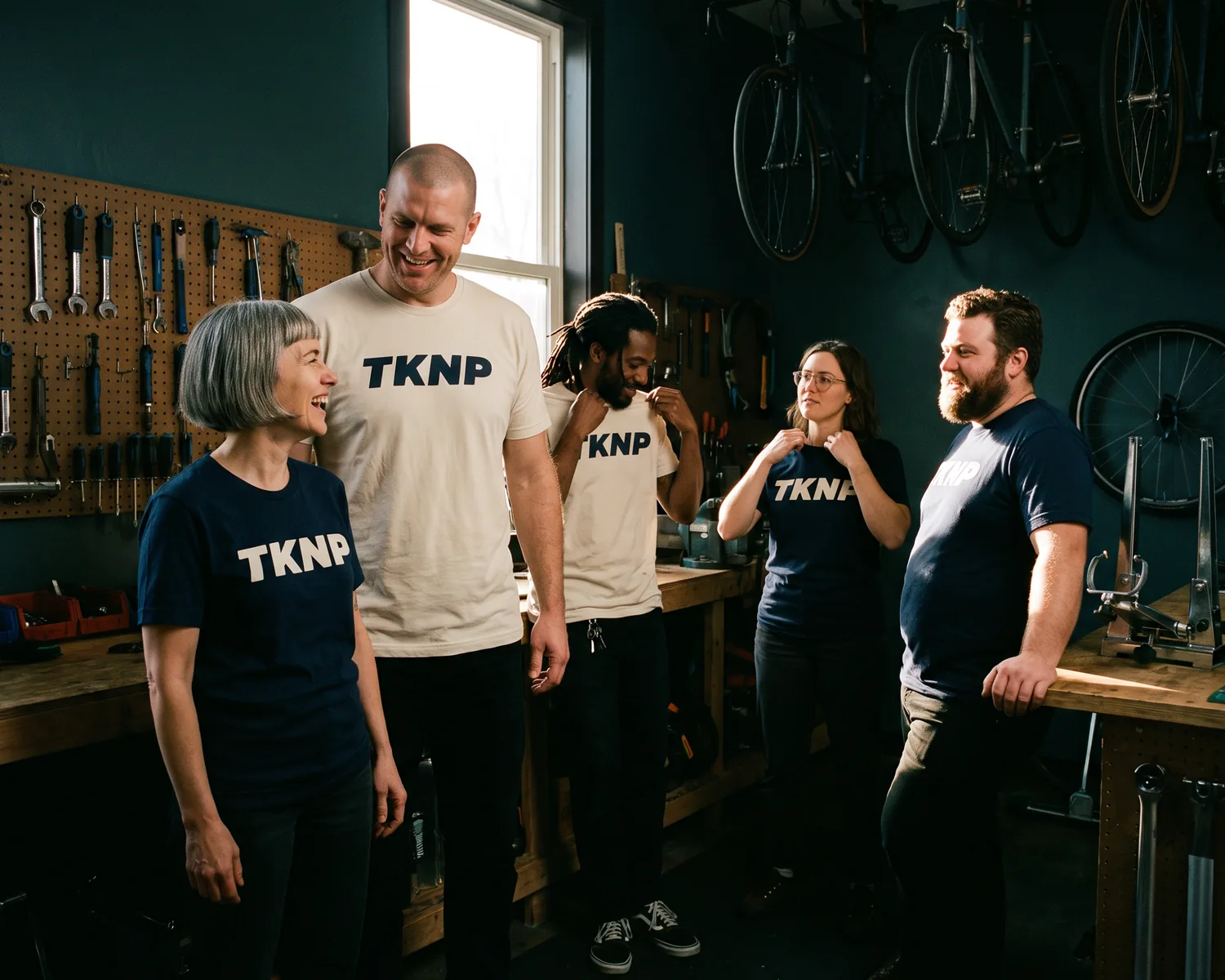 In a bike workshop, five team members, from size S to XL, wear the same custom TKNP t-shirt, in ecru and navy.
