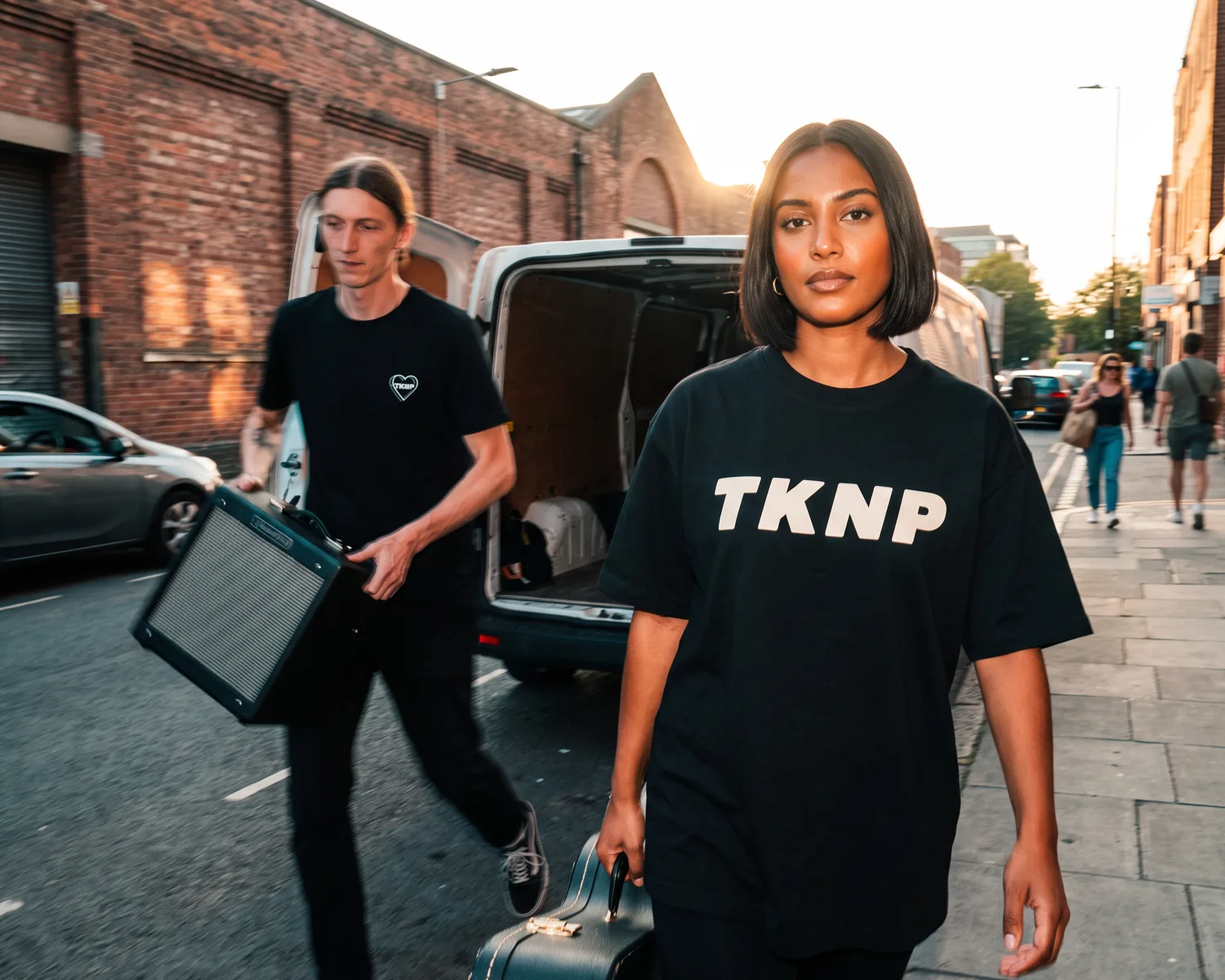 Outside a rehearsal space, a singer in a black t-shirt with a large screen-printed TKNP logo walks forward with a guitar case while a bassist in a black t-shirt with an embroidered heart logo loads the amp.