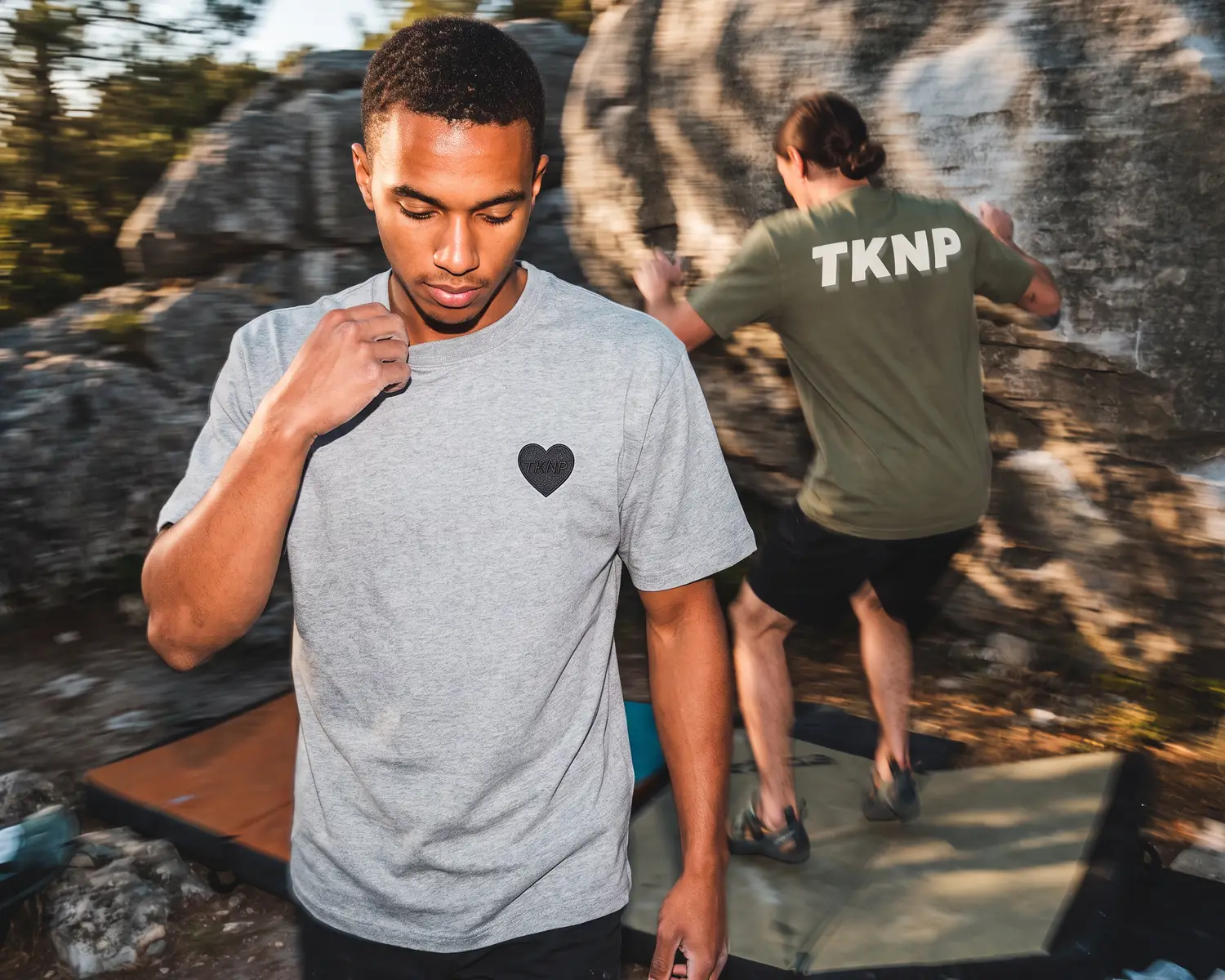 At the foot of the bouldering rocks, a climber straightens the collar of his heather grey t-shirt with a small embroidered TKNP heart logo, while another, in an olive t-shirt screen printed with TKNP on the back, jumps onto the crash pad.