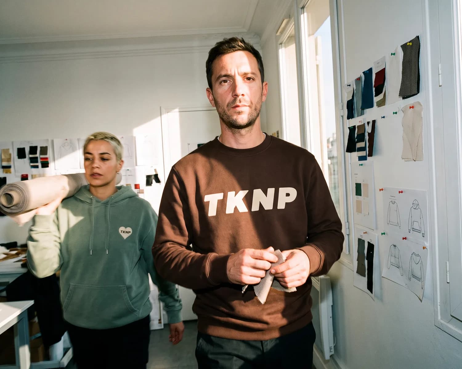 In a small brand's workshop, the founder, in a chocolate sweatshirt screen printed with TKNP, feels a fabric swatch while a colleague in a sage hoodie with an embroidered heart logo walks past carrying a roll of fabric.
