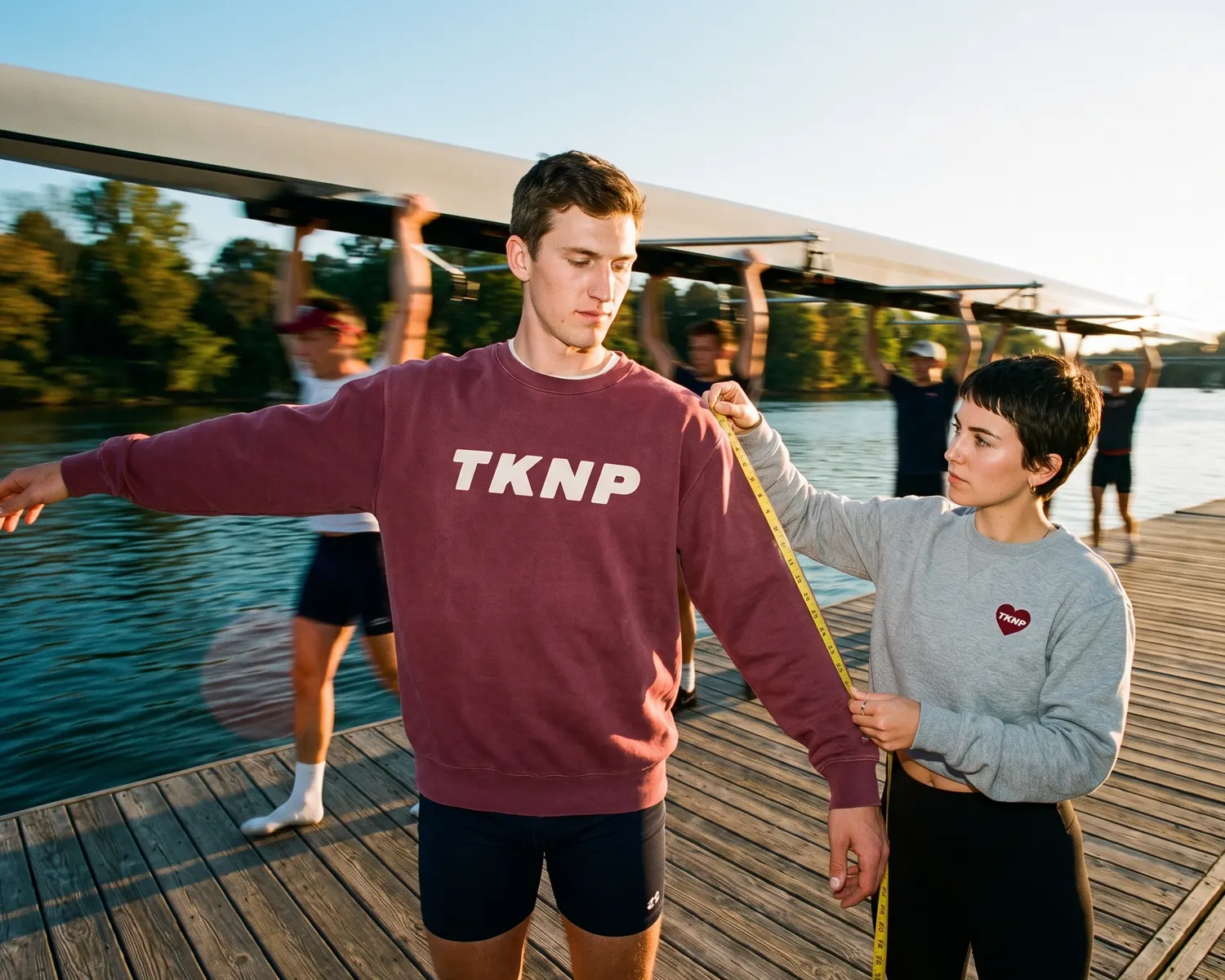 On a rowing club's dock, a teammate measures the sleeve of a rower's burgundy sweatshirt screen printed with TKNP with a tape measure, while the crew carries the boat past behind them.