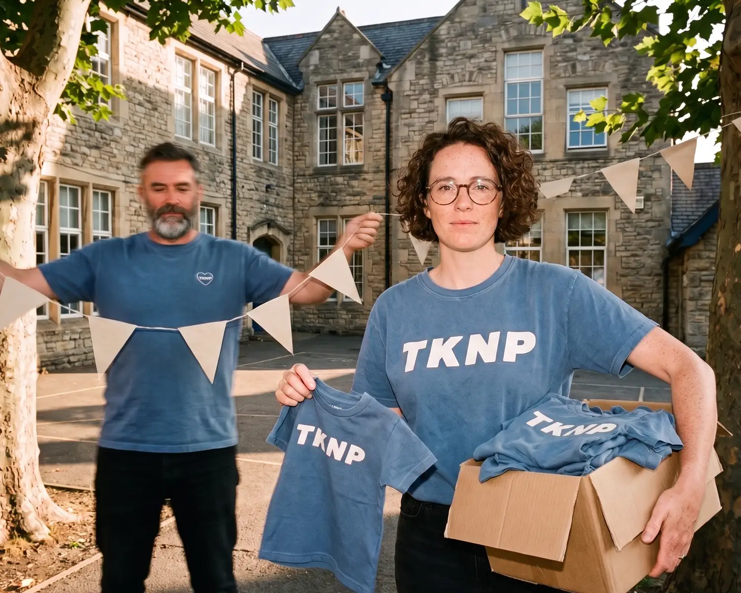 On the morning of the school fair, in the primary school courtyard, a teacher in a washed blue custom TKNP t-shirt brings a box of custom kids' t-shirts while a parent volunteer hangs the bunting.