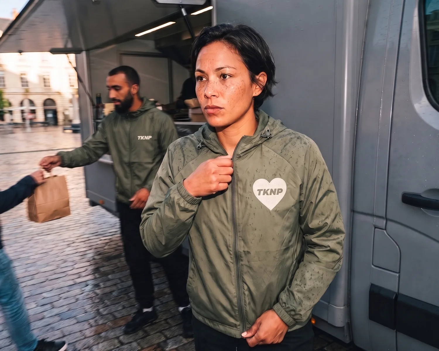 By a food truck on a square still wet from the rain, a cook zips up her olive polyamide windbreaker with raindrops beading on it and an embroidered TKNP heart logo on the left chest, while a colleague in an olive windbreaker with a small TKNP logo se