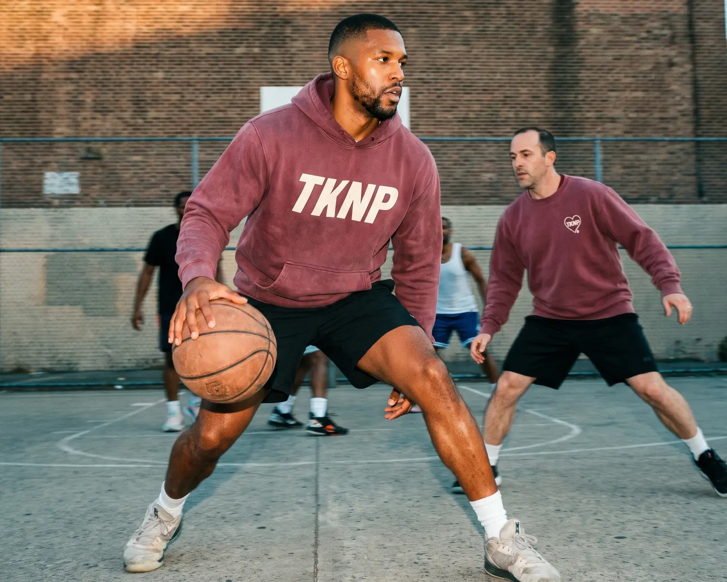 On a basketball club's outdoor court, a player in a custom burgundy hoodie with a large screen-printed TKNP logo dribbles past a teammate in a burgundy crew neck sweatshirt with an embroidered heart logo.