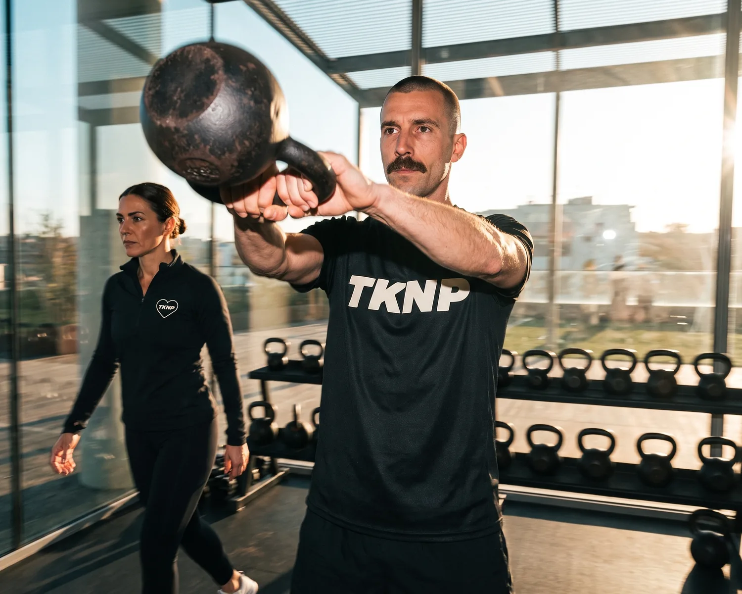 Dans une salle de fitness baignée de soleil, un adhérent en t-shirt de sport noir en polyester au grand logo TKNP sérigraphié lance un kettlebell, pendant que la coach, en legging noir et haut zippé au logo cœur TKNP brodé, passe derrière.