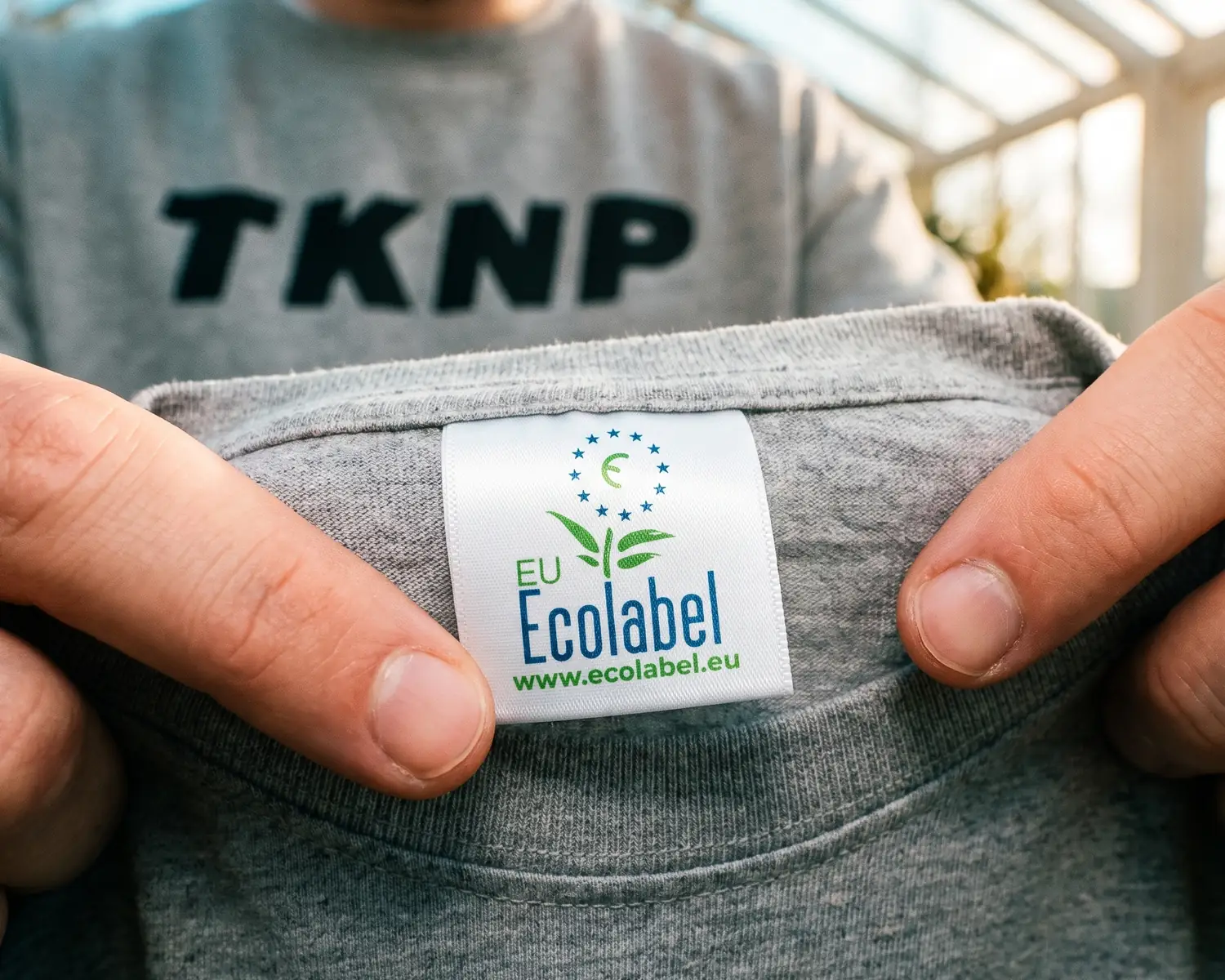 Étiquette Écolabel européen : la fleur de l'EU Ecolabel cousue au col d'un t-shirt vierge gris chiné, tenue en gros plan devant un sweat au grand logo TKNP.