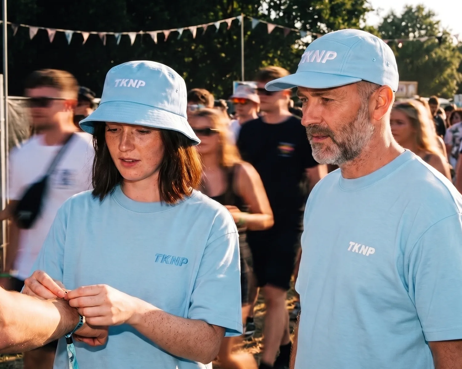 Bob ou casquette personnalisés : à l'entrée d'un festival, une bénévole en bob bleu ciel brodé TKNP, son t-shirt brodé ton sur ton côté cœur, et son collègue en casquette bleu ciel brodée du même logo accueillent le public.