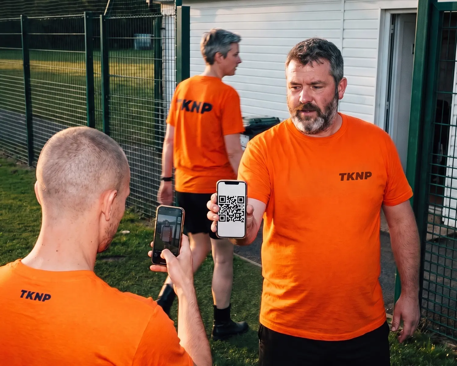 Prévente de t-shirts avec un QR code : au bord du terrain, un bénévole du club tend son téléphone, qui affiche le QR code de la campagne, à un joueur ; son t-shirt orange porte le logo TKNP sérigraphié côté cœur, dans un orange plus foncé.