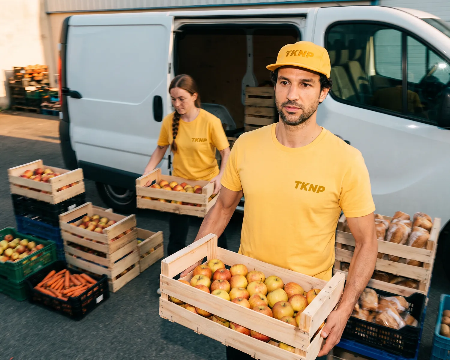 T-shirt d'association personnalisé pour les bénévoles : un bénévole en t-shirt et casquette jaune soleil, logo TKNP sérigraphié côté cœur dans un jaune plus foncé, porte une caisse de pommes vers la camionnette d'une banque alimentaire.