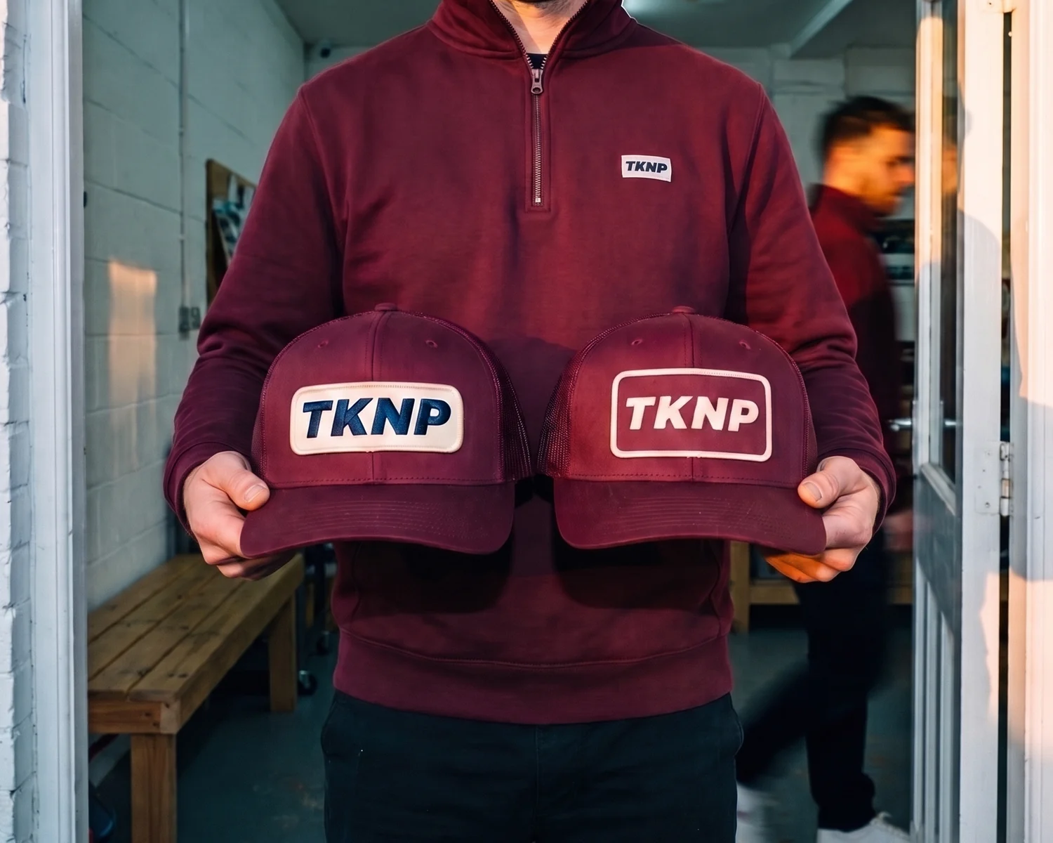 Écusson brodé ou tissé : deux casquettes trucker bordeaux tenues côte à côte, l'une avec un écusson brodé TKNP en relief sur fond crème, l'autre avec un écusson tissé TKNP plat sur fond bordeaux.