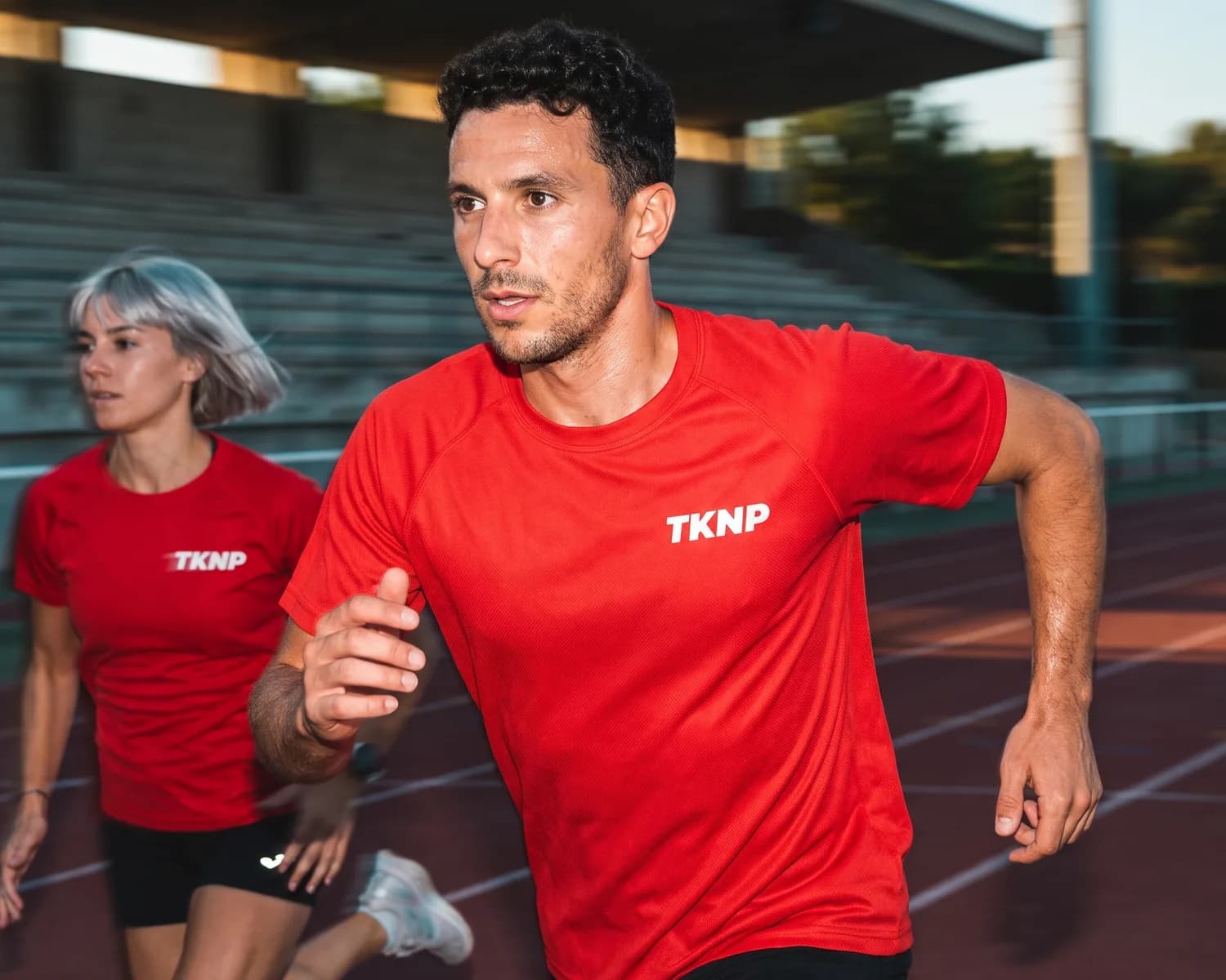 Impression textile polyester : un coureur en t-shirt technique rouge en maille polyester, le logo TKNP en transfert blanc net et plat côté cœur, pendant une séance sur piste.