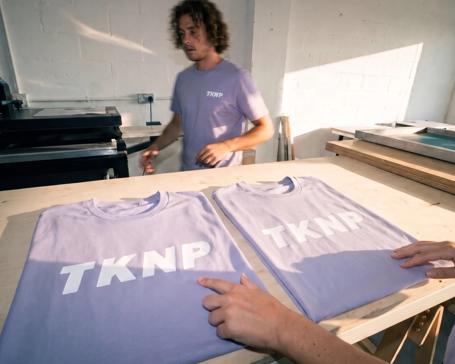DTF ou sérigraphie : deux t-shirts lavande posés côte à côte dans un atelier, le logo TKNP en sérigraphie blanche mate à gauche et en transfert DTF blanc au léger brillant à droite, comparés à la main.