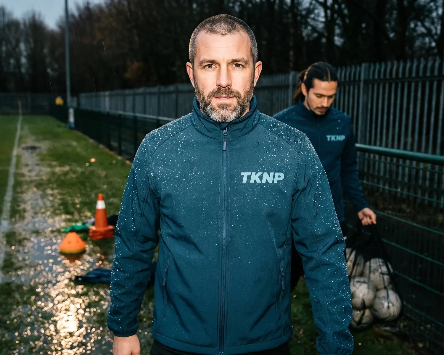 Veste déperlante bleu pétrole : l'eau perle en gouttes sur l'épaule et la manche d'un entraîneur au bord d'un terrain mouillé, logo TKNP imprimé ton sur ton côté cœur.