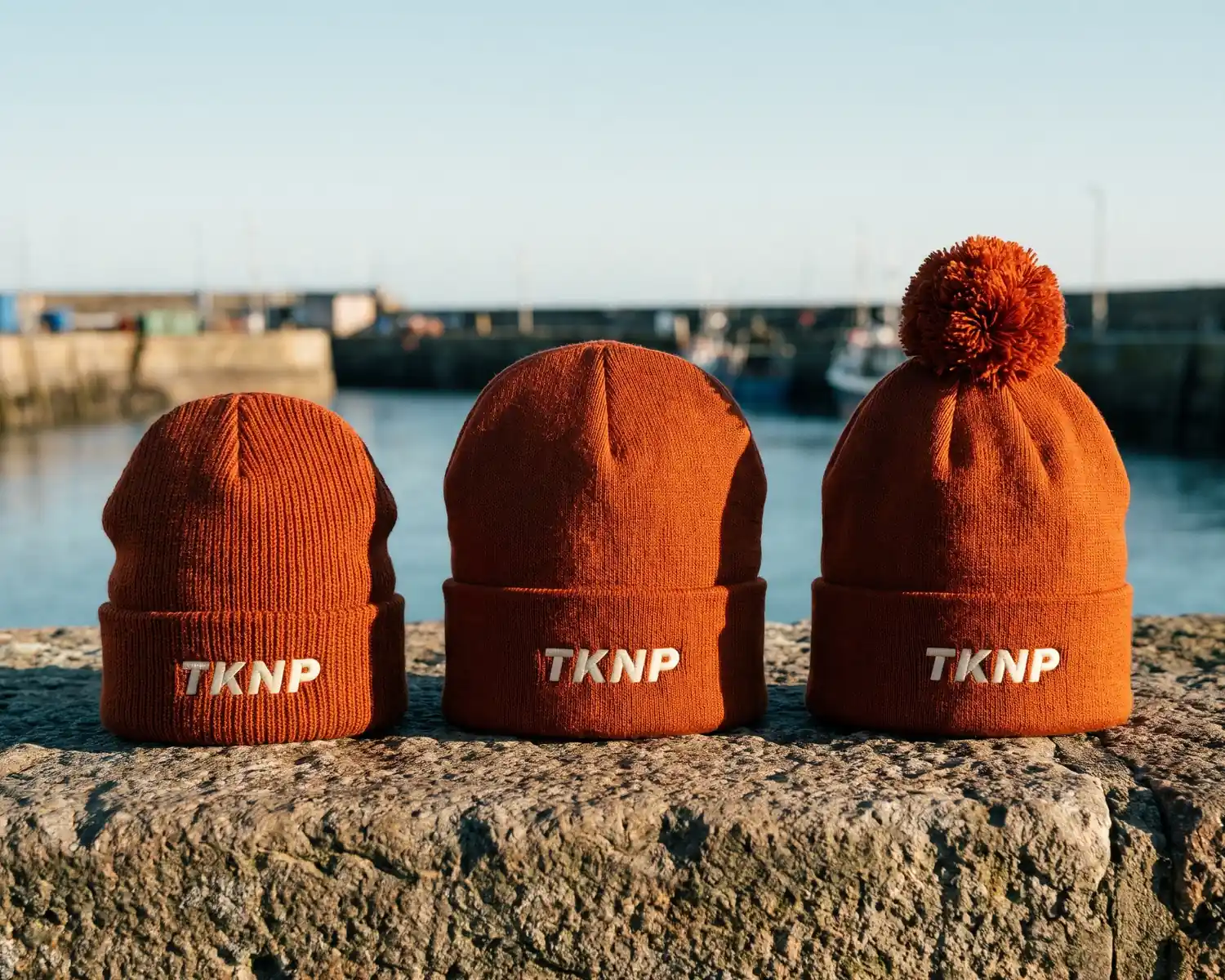 Types de bonnets personnalisés : un bonnet pêcheur côtelé, un bonnet à revers et un bonnet à pompon rouille posés côte à côte sur un muret de port, chacun brodé du logo TKNP en écru sur le revers.